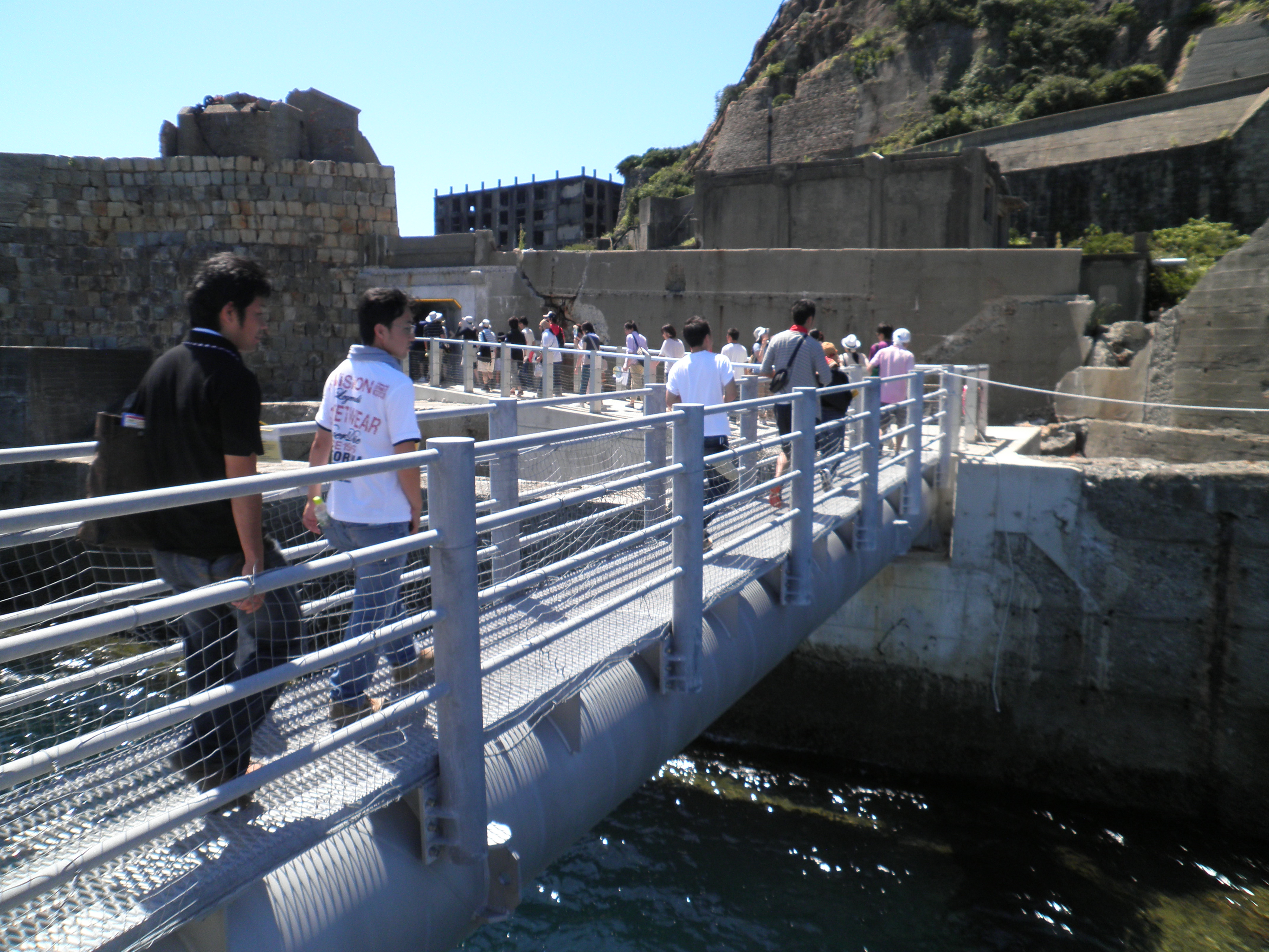 Tourists walking across a metal gangway to land on Hashima Island, with deteriorating concrete seawalls and abandoned apartment buildings visible in the background